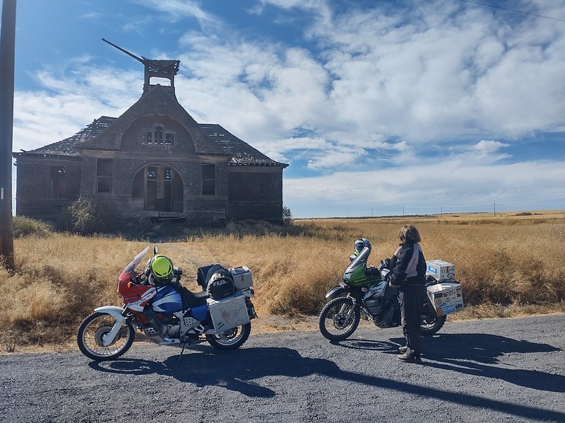two motorcycles in front of an old schoolhouse that is falling apart. A woman stands looking at it with her back to the camera.