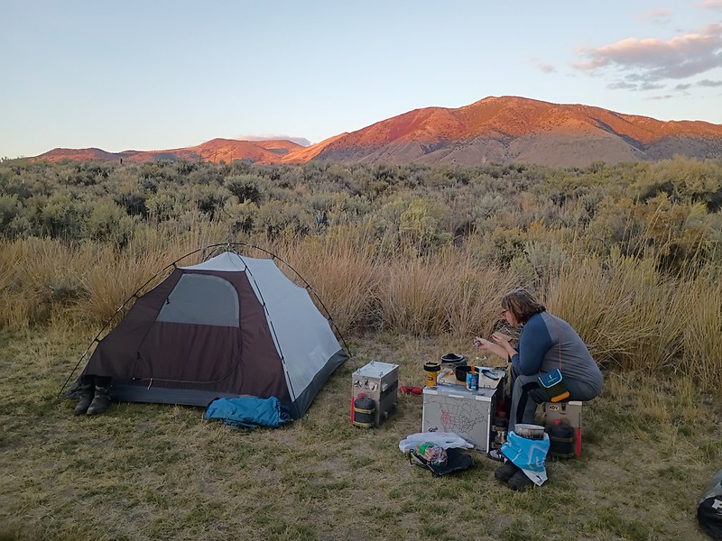 a woman sits on a
        motorcycle pannier at a table made from other panniers next to a
        tent in the middle of a field