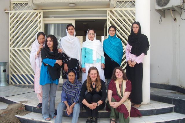 a group of women stand or sit in front of the doorways of an office. Many are in head scarves. Some smile, some have blank faces. There are many stories on the faces in this photo.