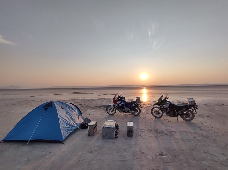 two motorcycles in the desert
        at sunrise with panniers as table and chairs next to a tent