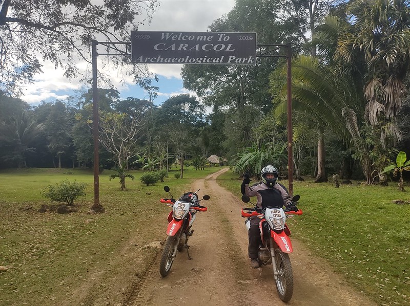 A woman on a motorcycle
next to a riderless motorcycle, under a sign in a jungle
regarding the ruins of Caracol in Belize