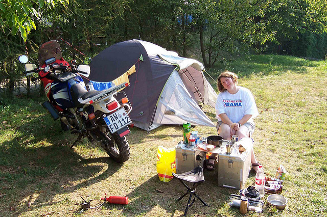 a woman in a
        "Texans for Obama" t-shirt sits at a table made from
        motorcycle panniers, next to a motorcycle and in frnt of a tent.
        She's at a campsite.